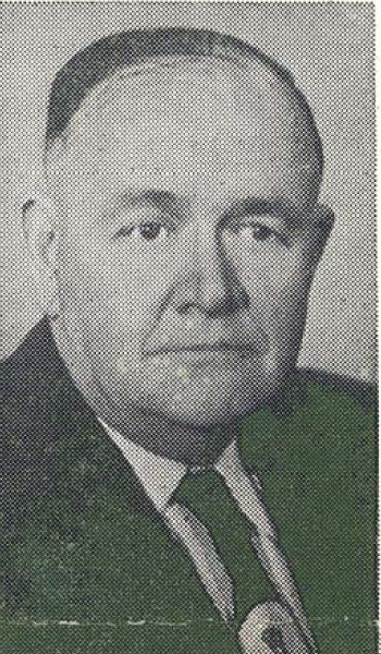 Black and white portrait of an older man with short hair, wearing a suit, white shirt, and tie, looking directly at the camera with a neutral expression.