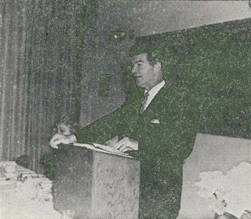 A man in a suit speaks at a podium in a room, possibly giving a lecture or presentation. Papers and objects are visible on the table beside him. The image appears to be in black and white.
