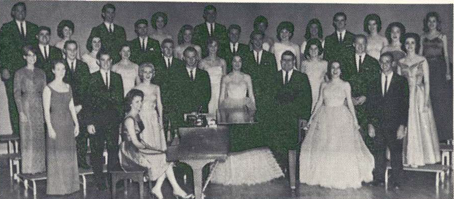 A black and white photo of a formal group of men and women standing and sitting in rows on a stage, some in suits and others in dresses, posed around a piano.