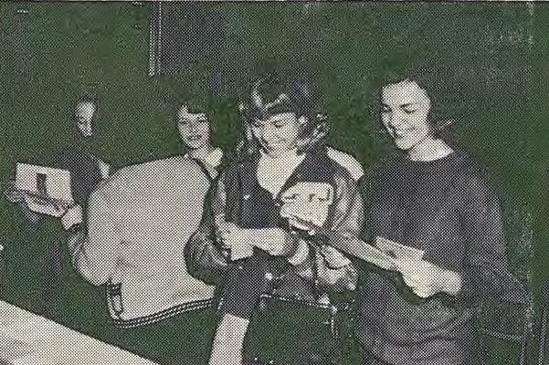 Four young women stand close together, looking down at papers they are holding. They appear focused and engaged, with a plain dark background behind them. The image is in black and white and has a grainy texture.