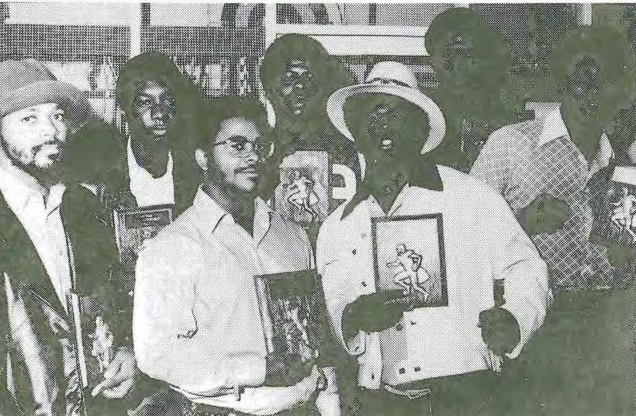 A group of men posing together indoors, each holding books or framed pictures. They are wearing hats and a variety of shirts, and appear to be smiling or talking to the camera. The image is in black and white.