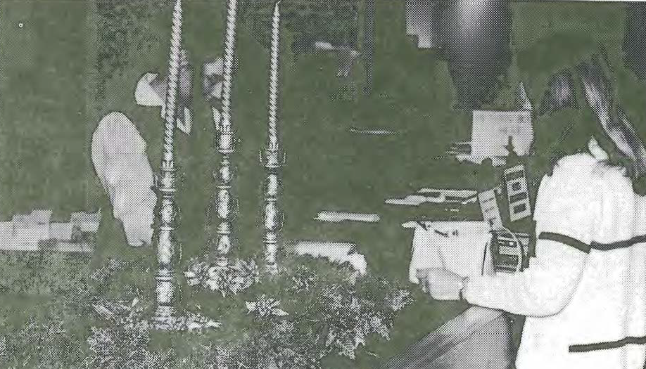 A woman stands at a counter in front of two tall candlesticks and greenery, interacting with a person on the other side who is partially obscured. Documents and office supplies are visible on the counter.