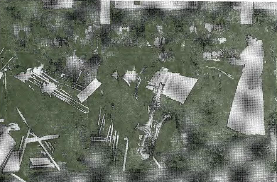 A black-and-white photo shows musicians playing brass instruments in an orchestra, with a woman in a light-colored dress standing and conducting them in front of music stands.