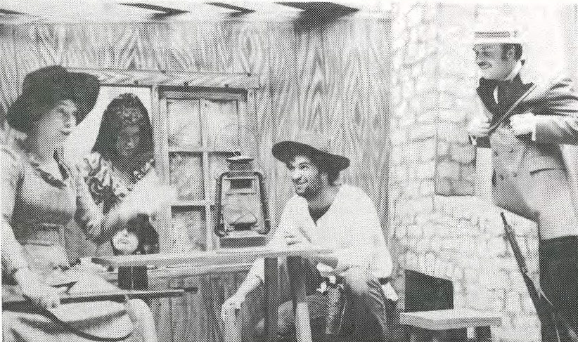 Four people in old-fashioned Western-style costumes sit and stand around a wooden table in a rustic room, smiling and conversing. The setting includes wooden walls and a stone fireplace.