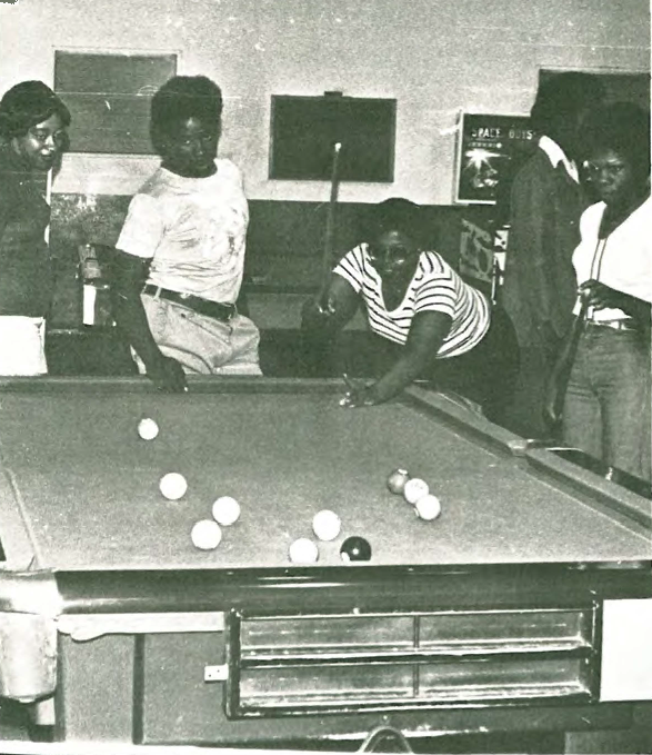 Four people stand around a pool table; one person leans over, preparing to take a shot, while the others watch. The table is scattered with balls. The scene appears to be in a recreational room.