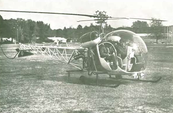 A vintage helicopter with an exposed metal frame and a bubble-shaped cockpit is parked on a grassy field, with trees and another aircraft visible in the background.