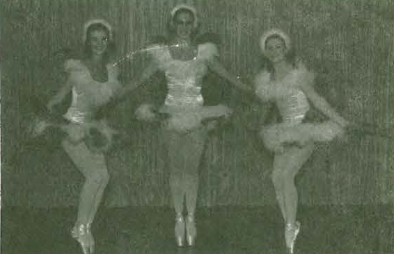 Three ballet dancers in tutus and headpieces pose together on stage, balancing on one leg with arms intertwined, against a plain backdrop. The image appears to be black and white or low contrast.