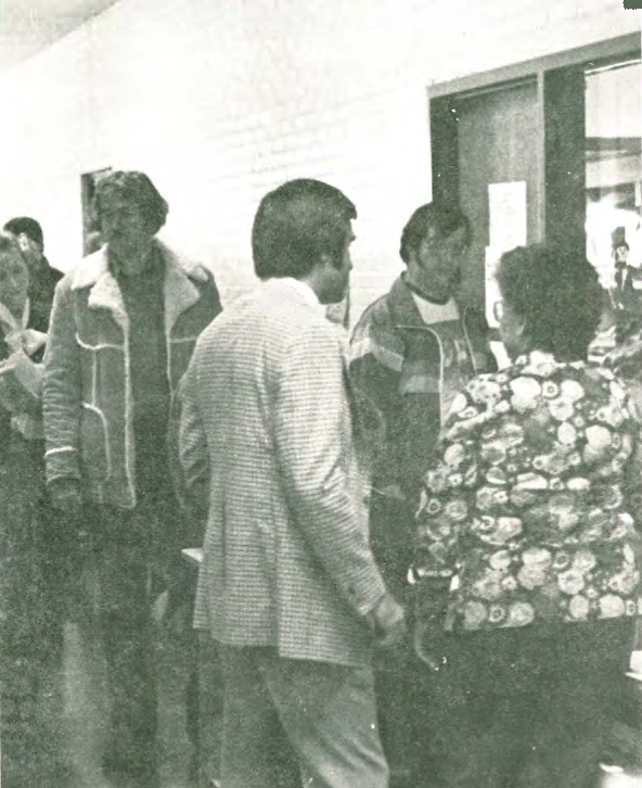 A group of people stand in a hallway near a windowed office door, wearing jackets and patterned clothing, engaging in conversation. The setting appears to be indoors, possibly in a school or community building.