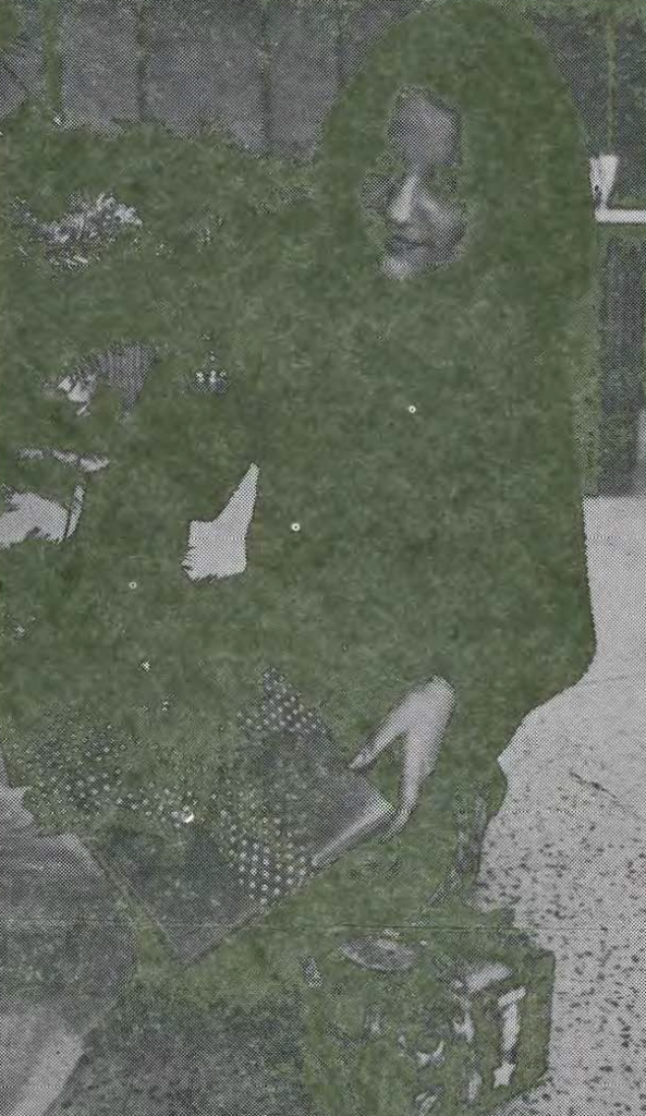 A person sits next to a decorated Christmas tree, smiling while holding an open box. Wrapped presents are visible nearby.