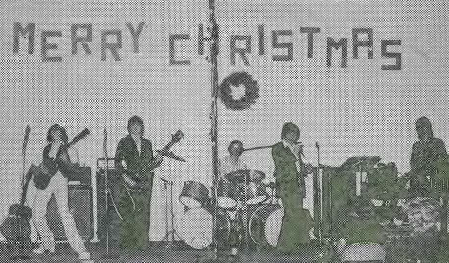 A black and white photo of a band performing on stage under a large MERRY CHRISTMAS sign with a wreath, featuring five musicians playing guitar, bass, drums, keyboard, and singing.