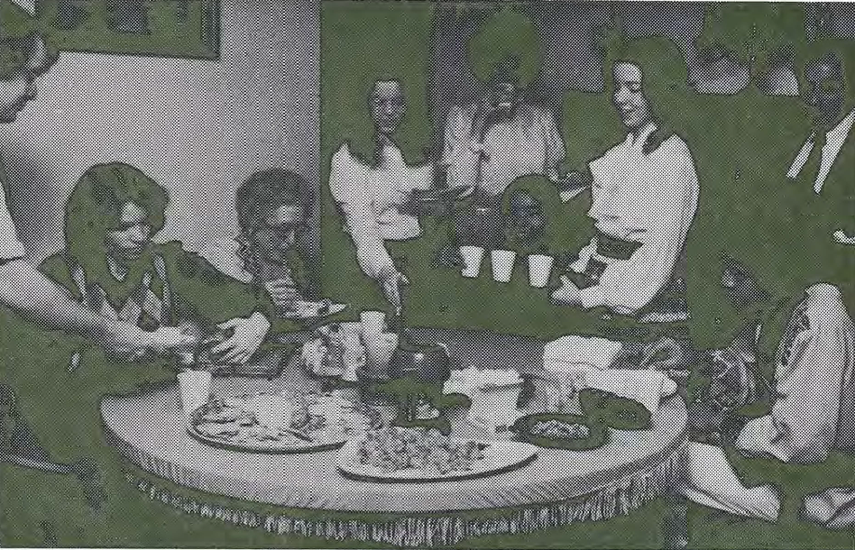 A group of people sit and stand around a round table, sharing food and drinks. The table is set with plates, cups, and a teapot. Some people are serving or pouring drinks, and everyone appears to be socializing.