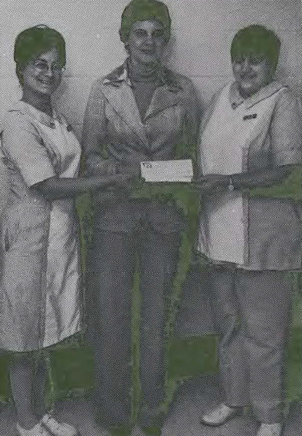 Three women stand together indoors, with two of them wearing uniforms and holding a rectangular object, possibly a check, between them. The woman in the middle is dressed in casual attire.