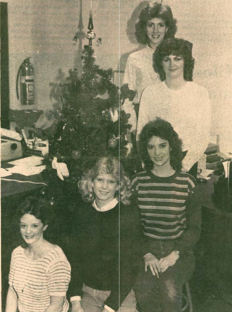 Five women pose indoors near a decorated Christmas tree, smiling at the camera. There is office equipment, paperwork, and a fire extinguisher visible in the background. The image appears to be from an earlier decade.