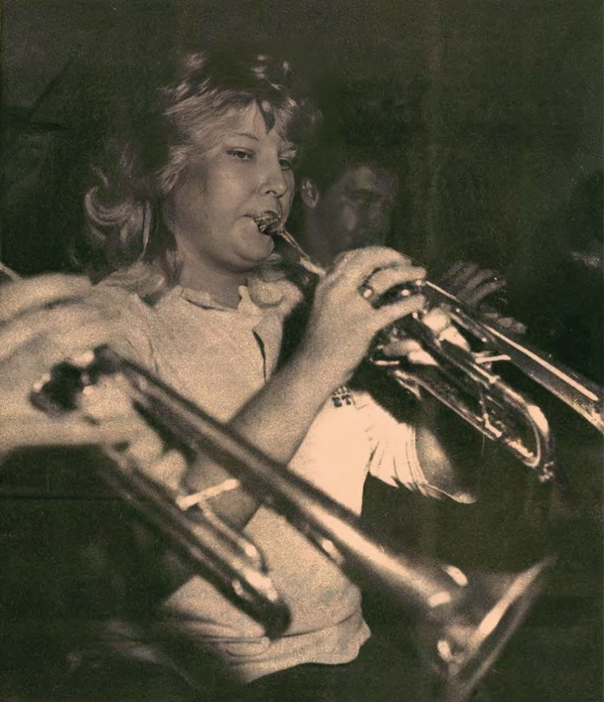 A woman plays a trumpet in a dimly lit room, with other musicians playing brass instruments in the background. She appears focused on her performance.