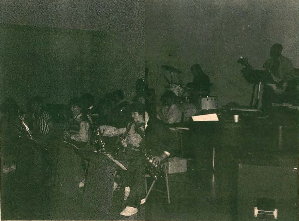 A dimly lit stage shows a group of musicians playing various instruments, including saxophones and drums, seated and performing together in a band or orchestra setting.