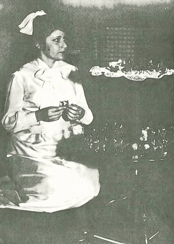 A woman in a white dress and headscarf sits, holding an object in her hands, with a table covered by a lace cloth and small items beside her; the setting appears vintage or historical.