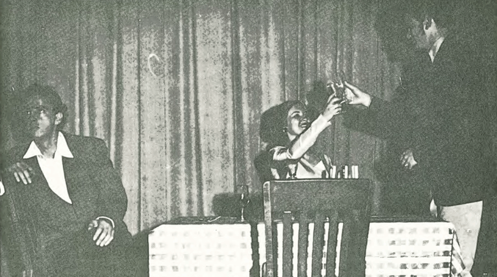 Three people sit at a table in front of a curtain. One person on the left sits alone, while the two on the right clink glasses in a toast. The scene appears to be from a performance or play.
