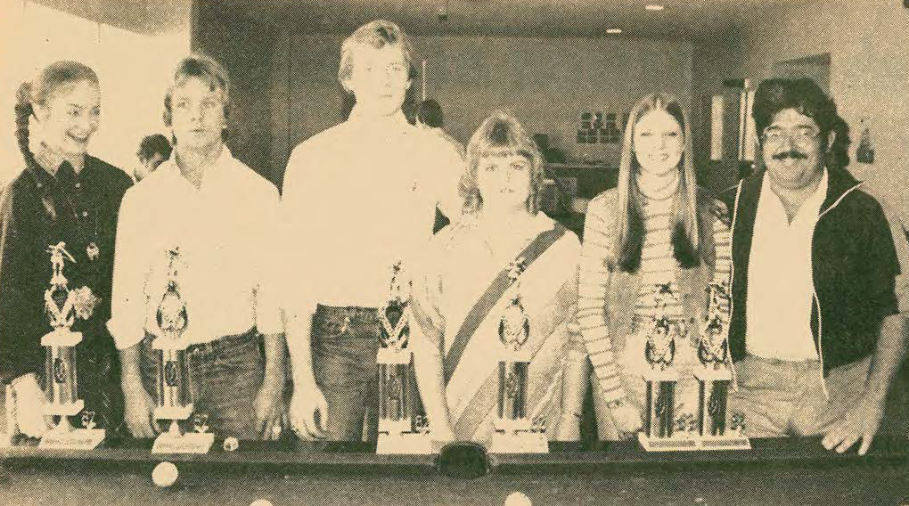 Six people stand behind a pool table displaying five large trophies. The group appears to be smiling or posing for the camera in an indoor setting, possibly celebrating a competition or tournament victory.
